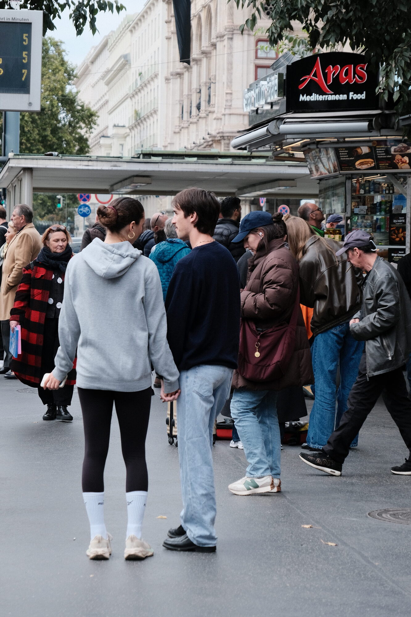 A couple holding hands at a tram stop wit ha Wurstel stand in the background
